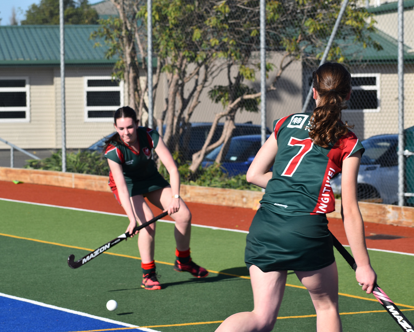 Two school aged girls playing hockey on a green and blue field. The girl in the backgroud is preparing to strike the ball.